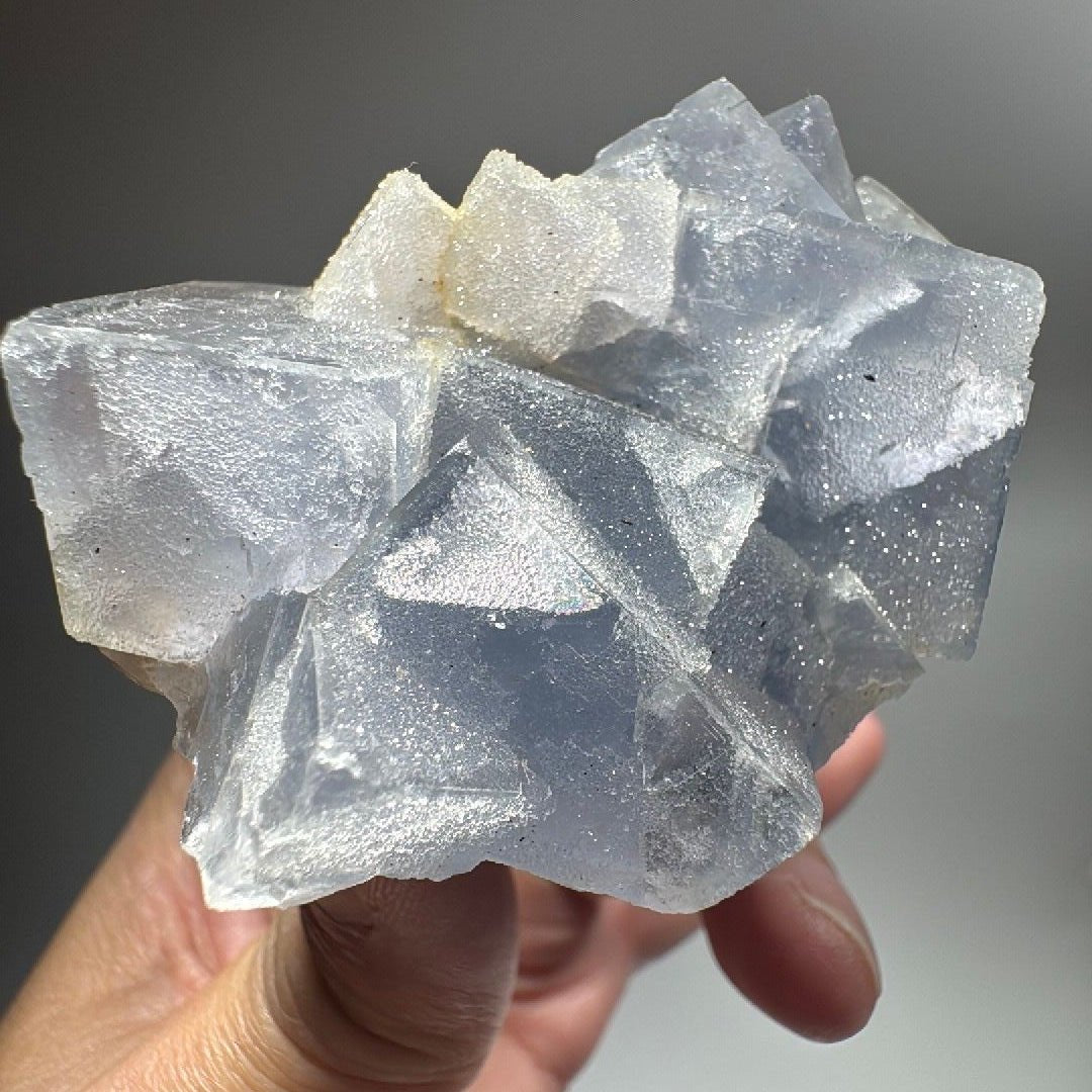 Hand holding a pale blue fluorite cluster, stepped cubic crystals frosted with sugary white quartz