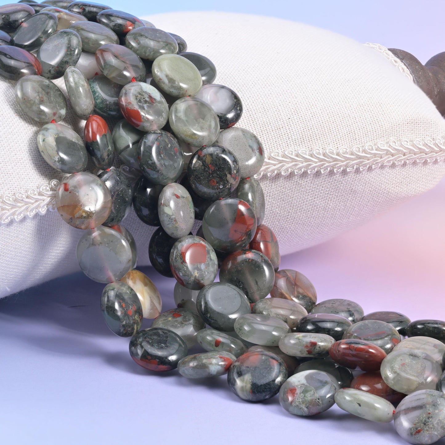 African Bloodstone coin strands spilling over a white cushion, grey-green flecked with red