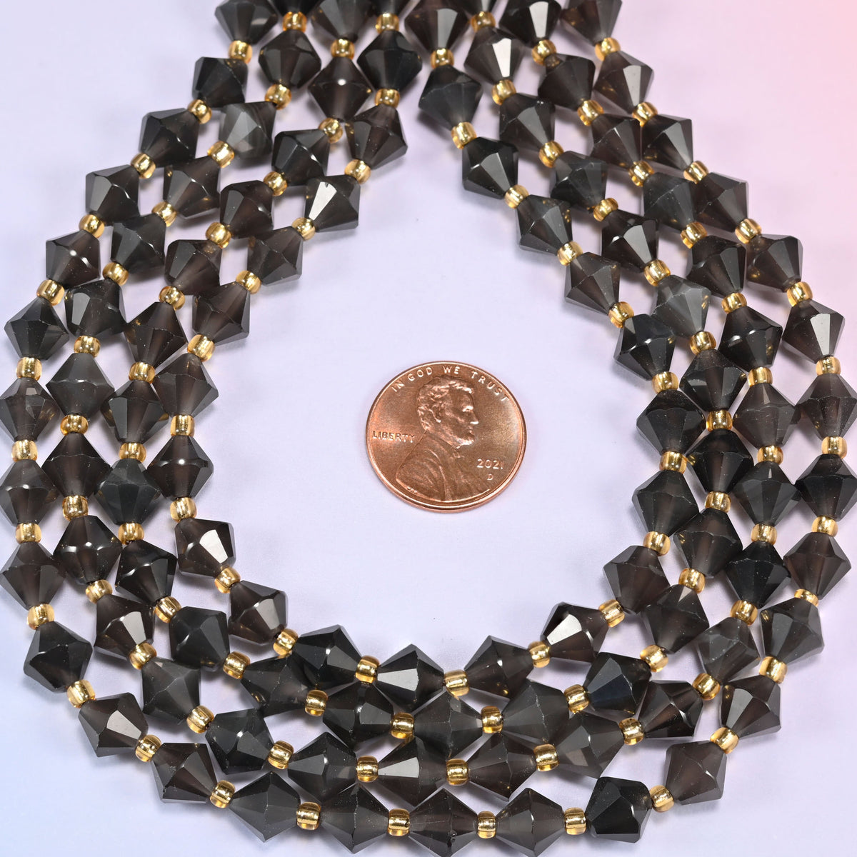 Ice Obsidian faceted bicone beads 8mm with gold-tone spacers, strand coiled into a neat circle with a penny centered for scale