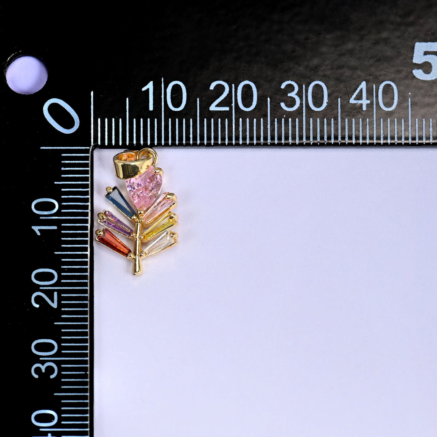 Multicolour leaf charm in gold with a pink stone and green, blue and orange baguette stones, beside a millimetre ruler for scale