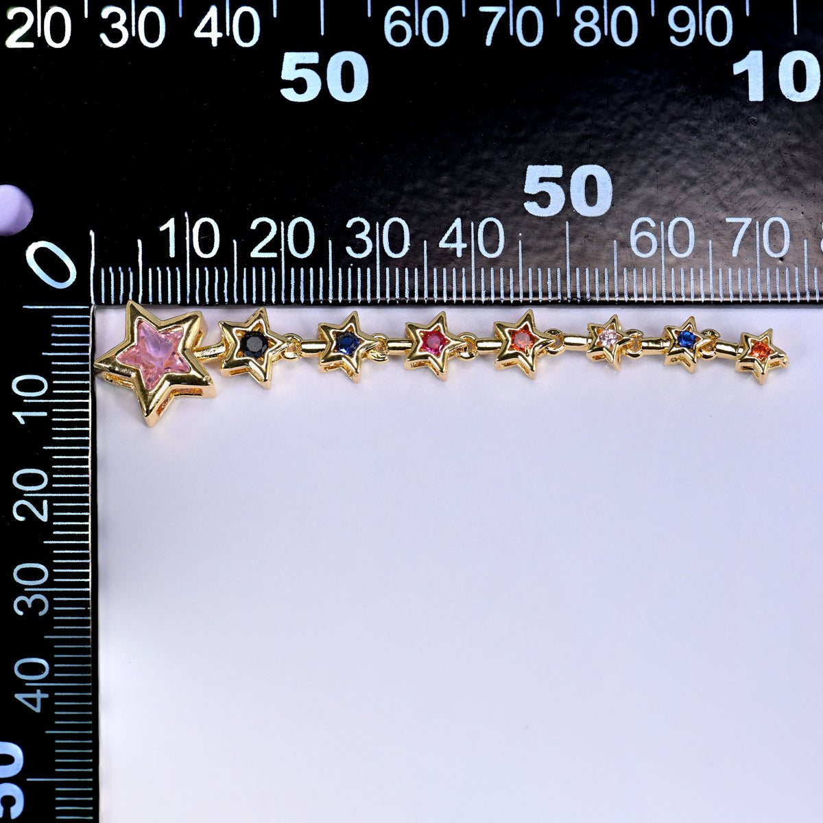 Shooting star pendant, a trail of small gold stars set with pink, blue and green stones leading to a larger pink star, laid along a millimetre ruler for scale
