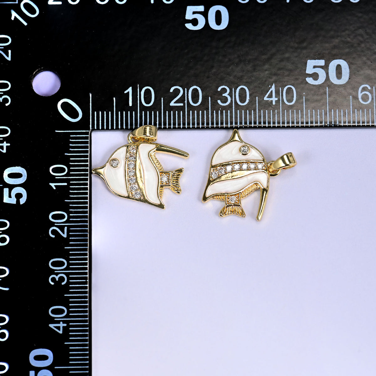 Two tropical fish charms in white enamel with gold fins and clear pave stones, shown front and angled, beside a millimetre ruler for scale