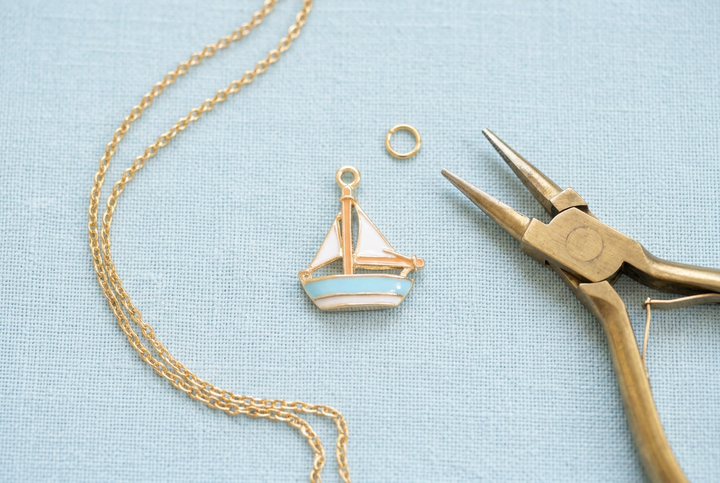 White and light blue enamel sailboat charm with gold-tone chain, jump ring, and jewelry pliers on pale blue linen