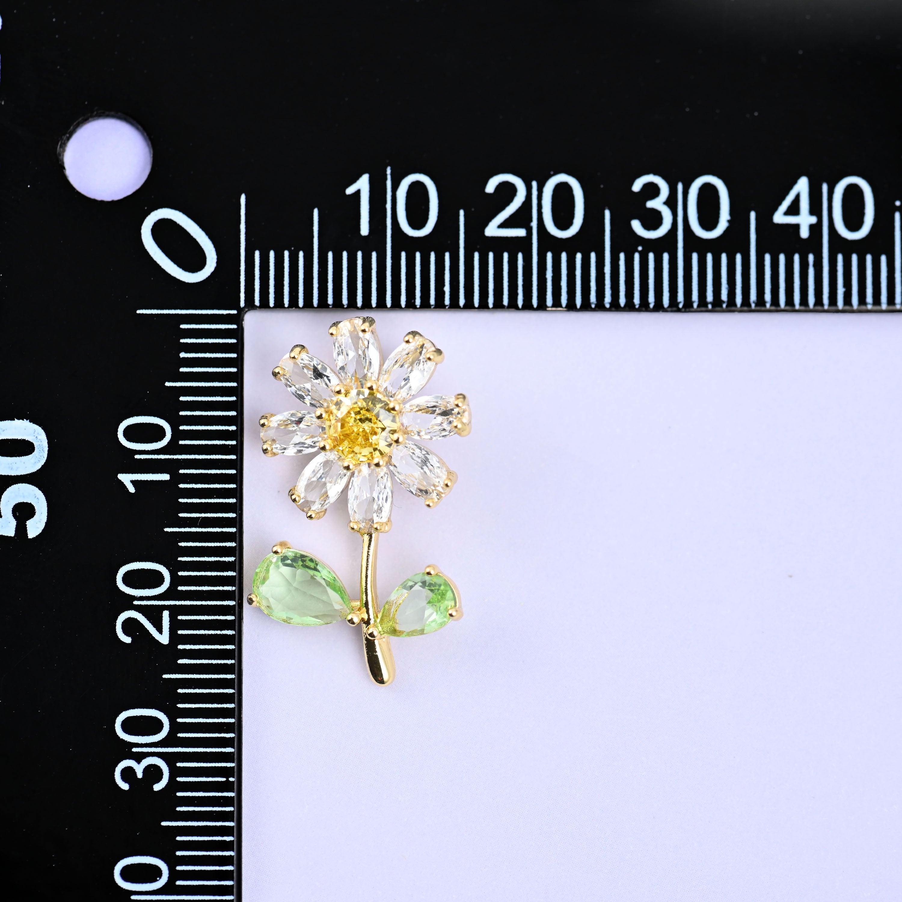 A daisy flower charm with clear petals, a yellow centre and green leaves, upright beside a millimetre ruler for scale