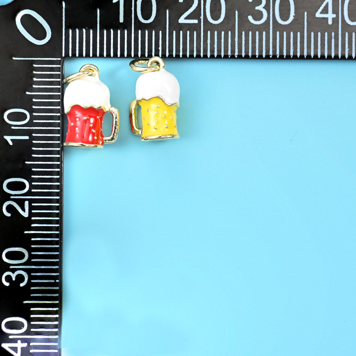Red and yellow enamel beer mug charms with white foam tops, beside a millimetre ruler for scale