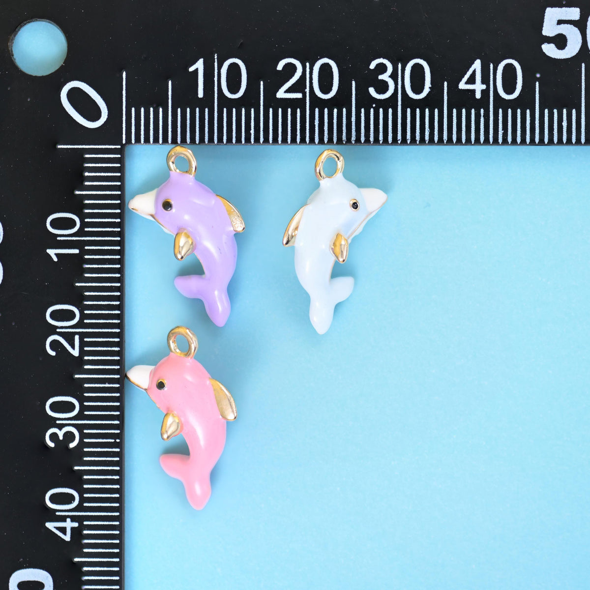 Lilac, white and pink enamel dolphin charms beside a millimetre ruler for scale