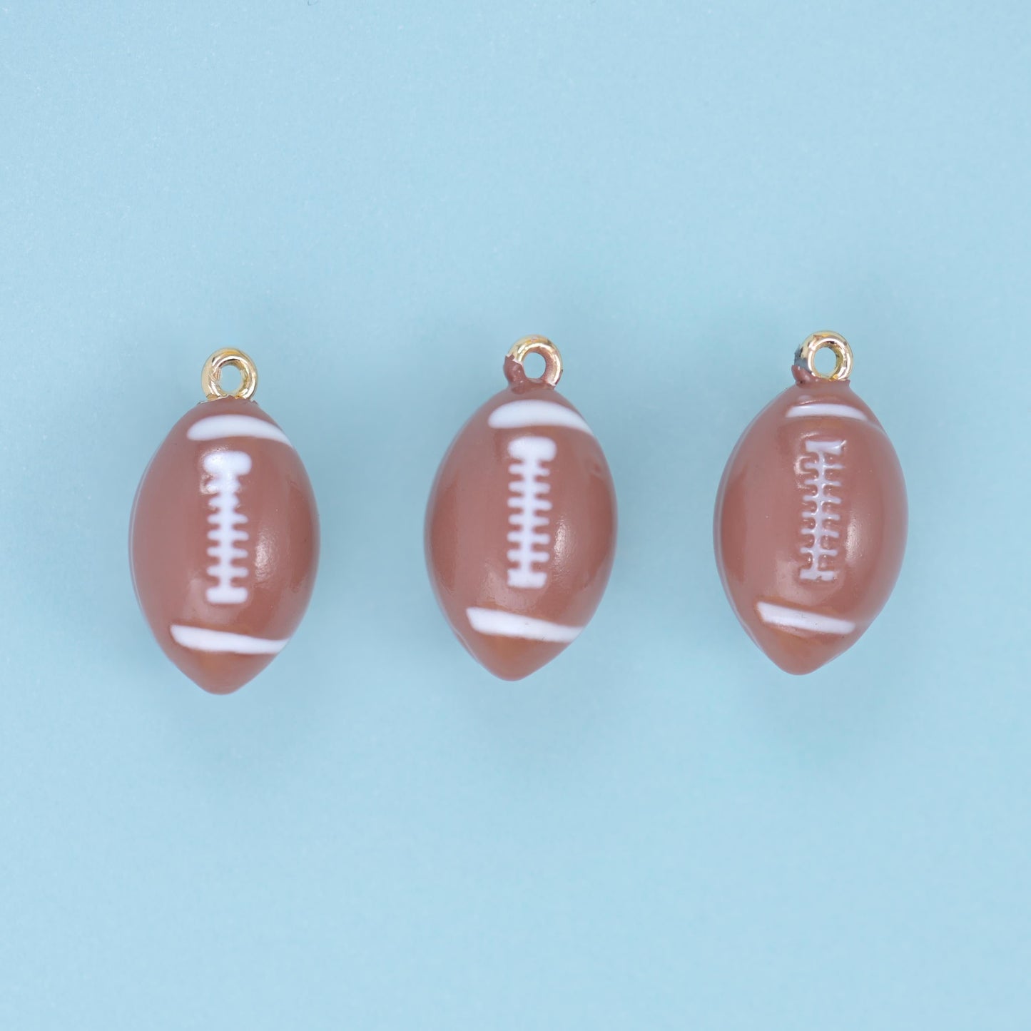 Three brown enamel football charms with white lacing lined up on a pale blue ground