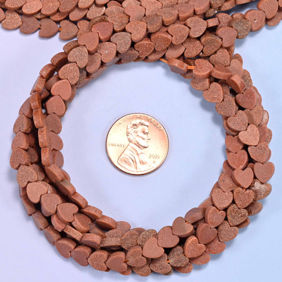 Matte Gold Sandstone flat heart bead strands coiled around a US penny for size reference