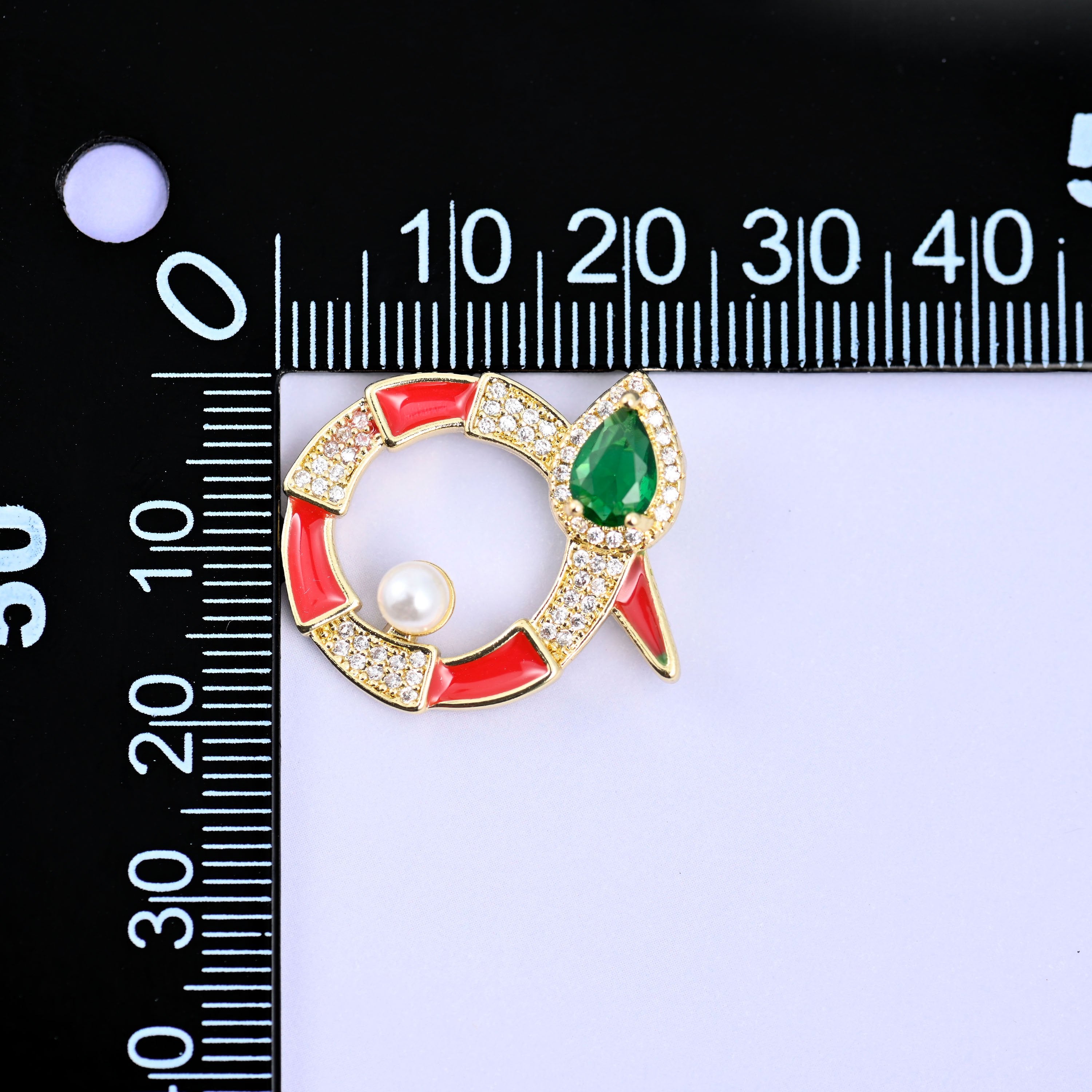 Round snake charm against a black millimetre ruler, the red enamel piece turned on its side with the green stone head pointing to the right.
