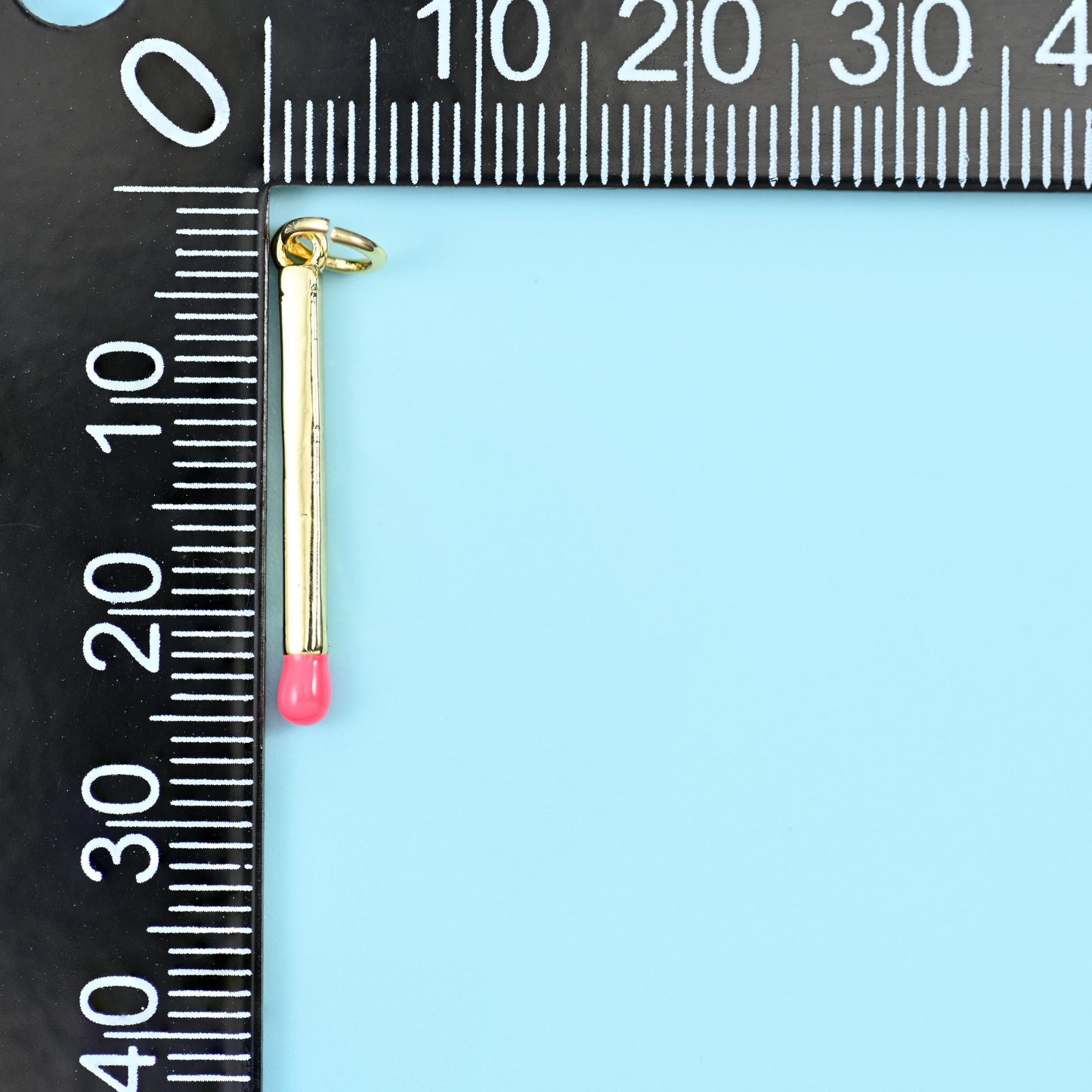 Matchstick pendant measured against a black millimetre ruler, the gold bar with a pink enamel tip running to about 23mm.