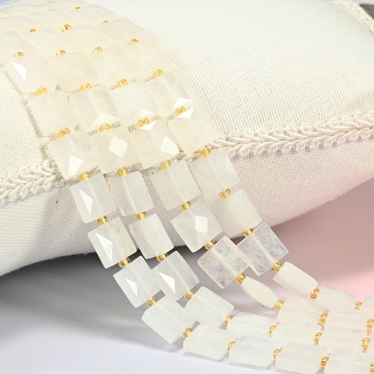 Clear quartz faceted flat square beads 10x10mm with small gold seed bead spacers, several strands draped over a white cushion