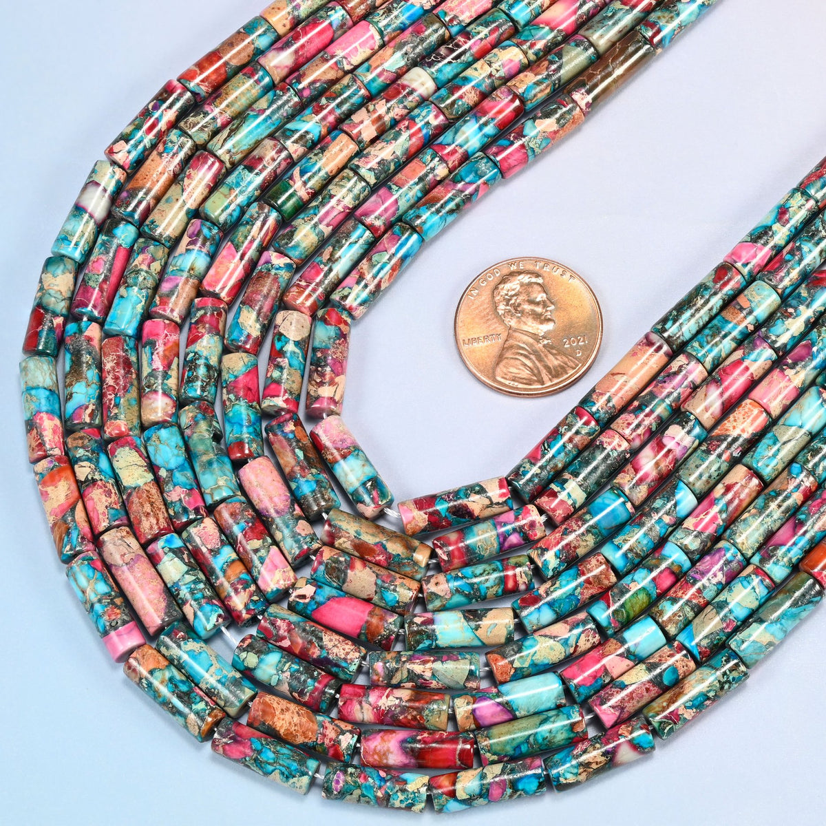 Close view of dyed sea sediment jasper tube bead strands curving beside a US penny for scale