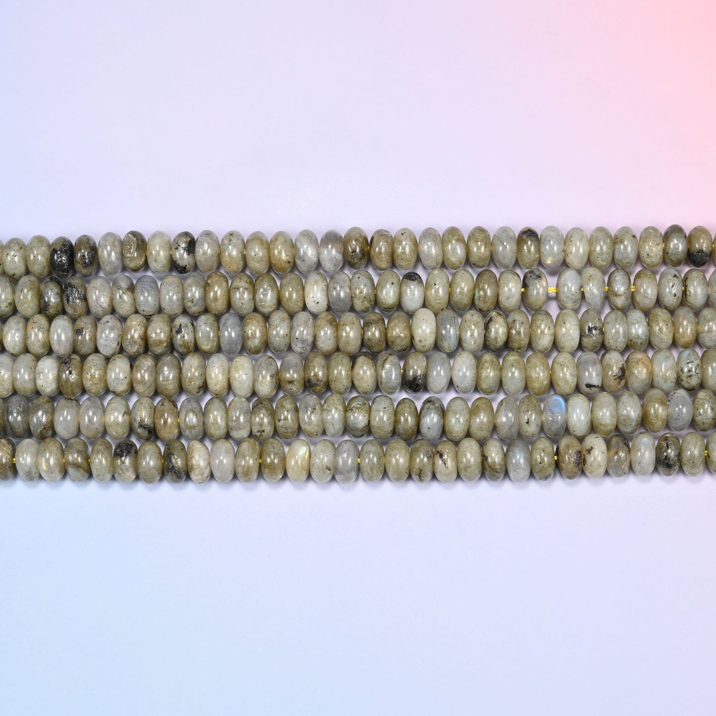 Overhead view of several rows of Grey Labradorite rondelle beads showing the soft grey and olive tones on a light lavender surface.