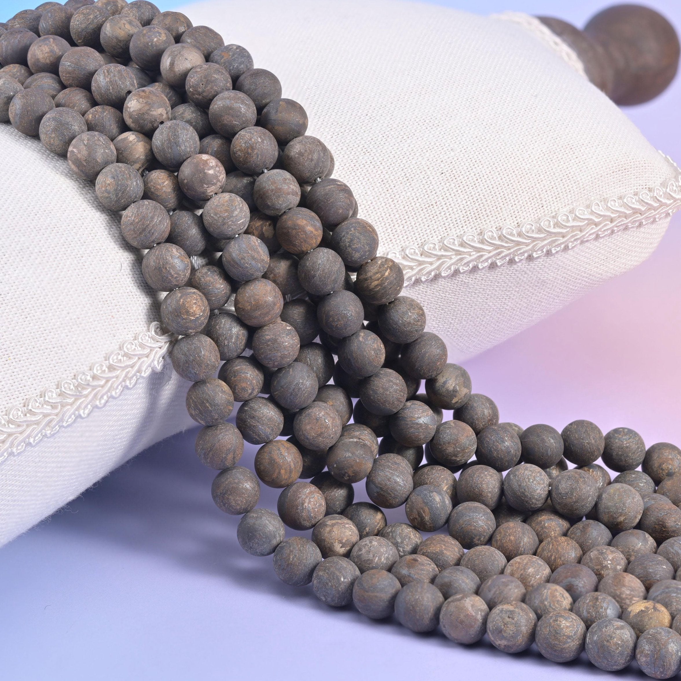 Matte bronzite round bead strands in mixed sizes resting against a white linen cushion, dry chalky surface with no shine