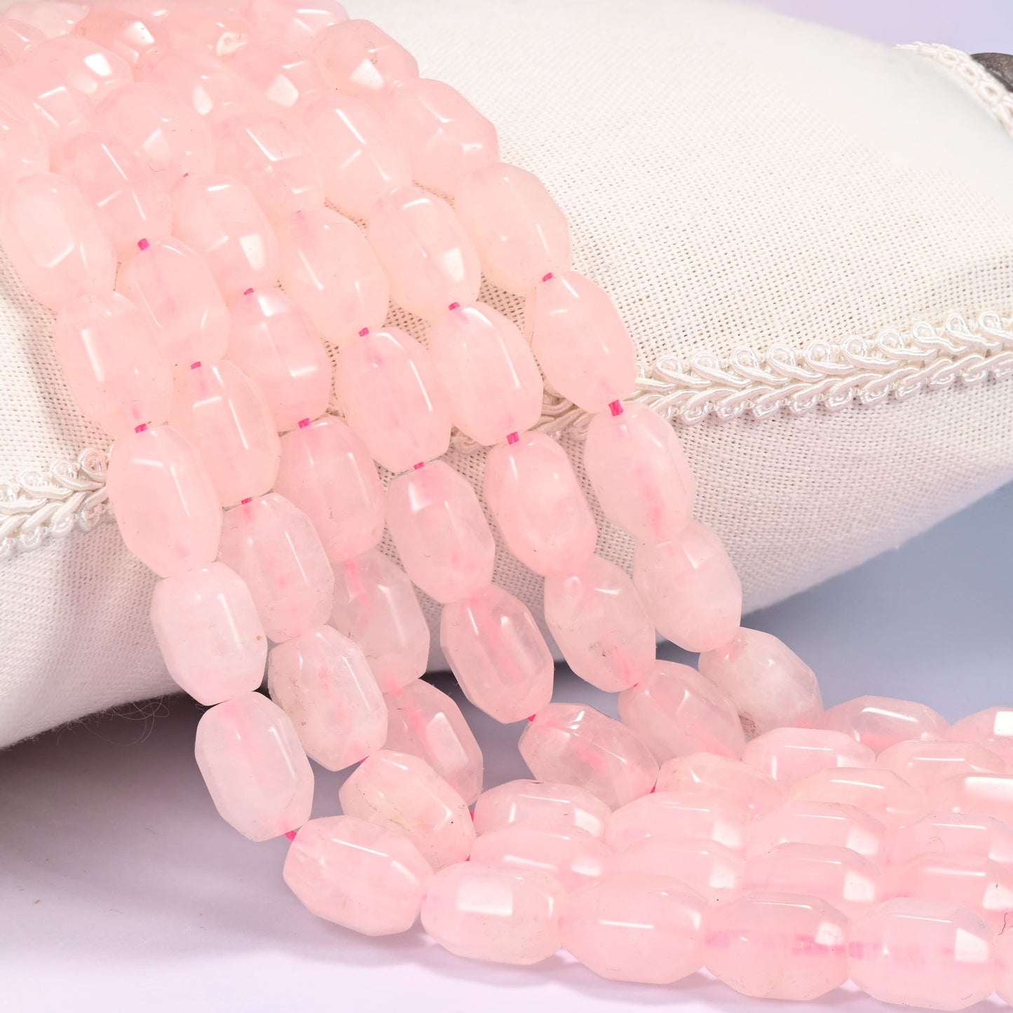 Close-up of multiple strands of soft pink faceted Rose Quartz barrel beads cascading diagonally over a cream woven pillow with decorative trim.