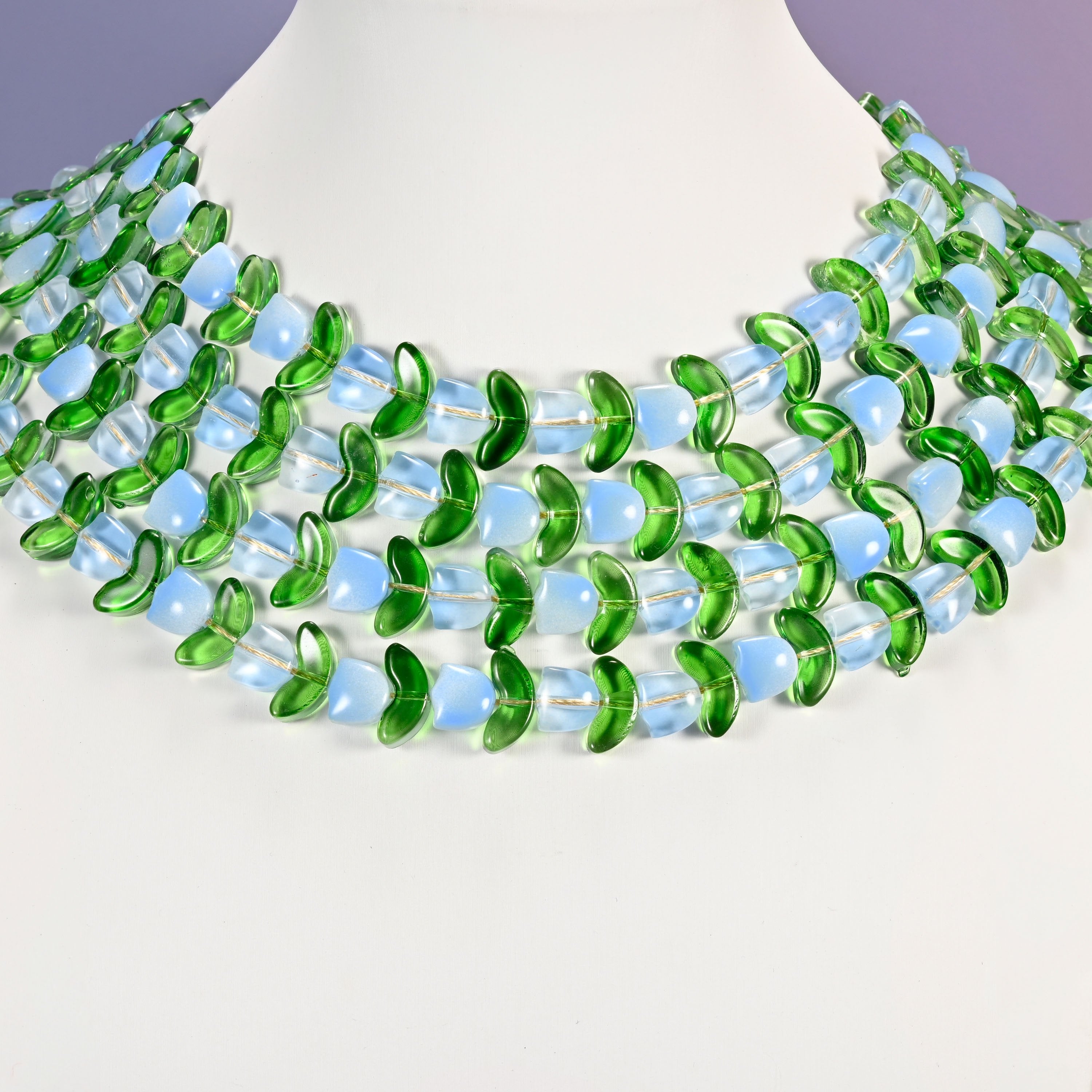 Multi-strand necklace of light blue tulip and green leaf glass beads laid on a white mannequin neck form against a lavender background.
