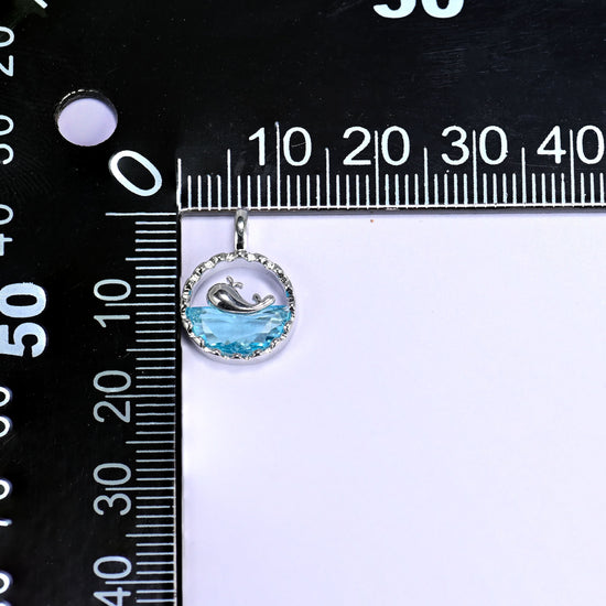 Platinum plated brass whale charm pendant with a silver whale silhouette diving into a faceted blue zirconia wave, set in a textured round frame.