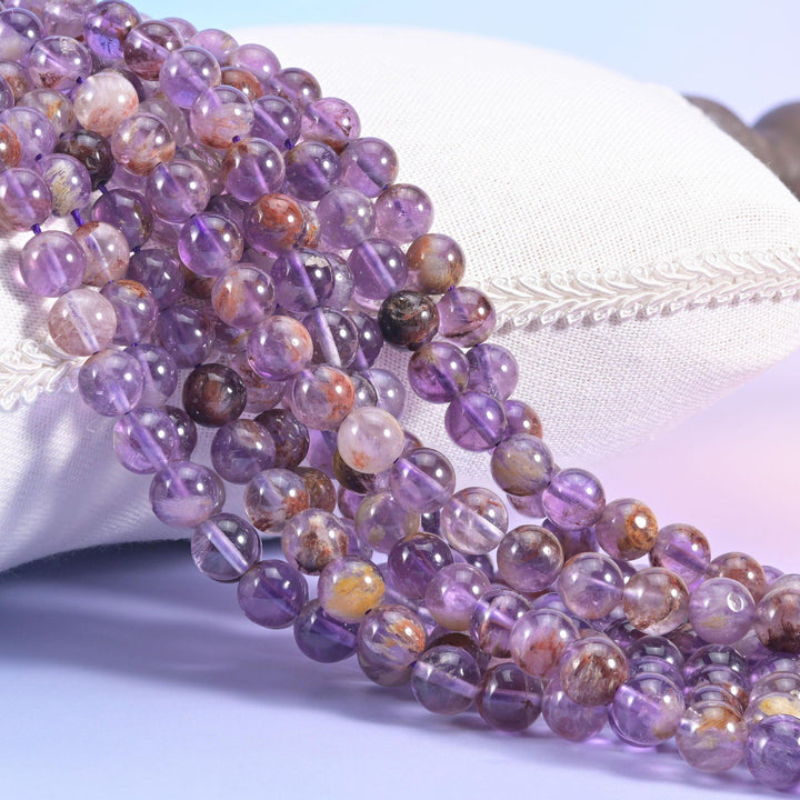 Super 7 Purple Phantom smooth round beads, strands draped over a white cushion, translucent lilac quartz with rusty red-brown inclusions