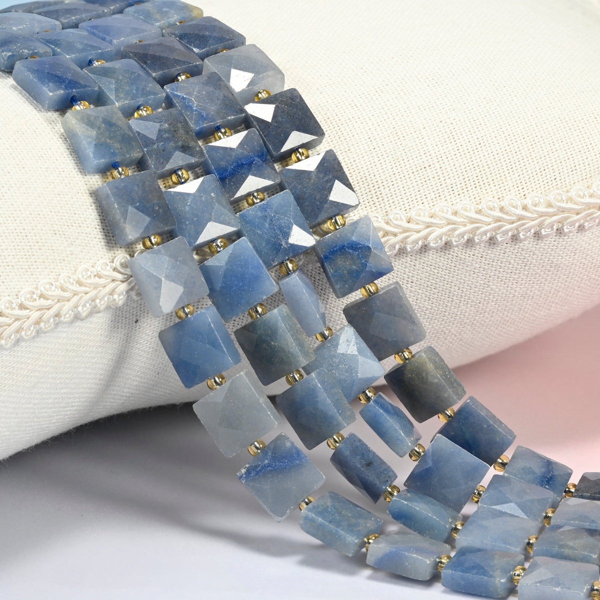 Natural blue aventurine faceted flat square beads 10x10mm with small gold seed bead spacers, several strands draped over a white cushion