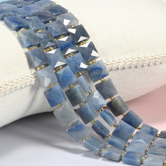 Natural blue aventurine faceted flat square beads 10x10mm with small gold seed bead spacers, several strands draped over a white cushion