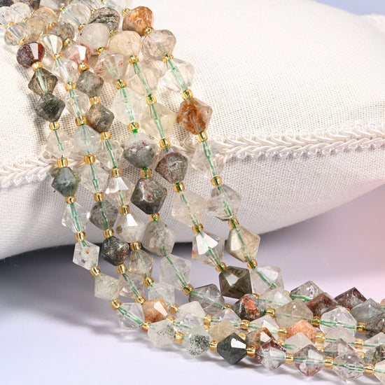 Green Phantom Quartz faceted bicone beads, 8mm and 10mm, draped diagonally over a white fabric cushion showing the internal green phantom inclusions.