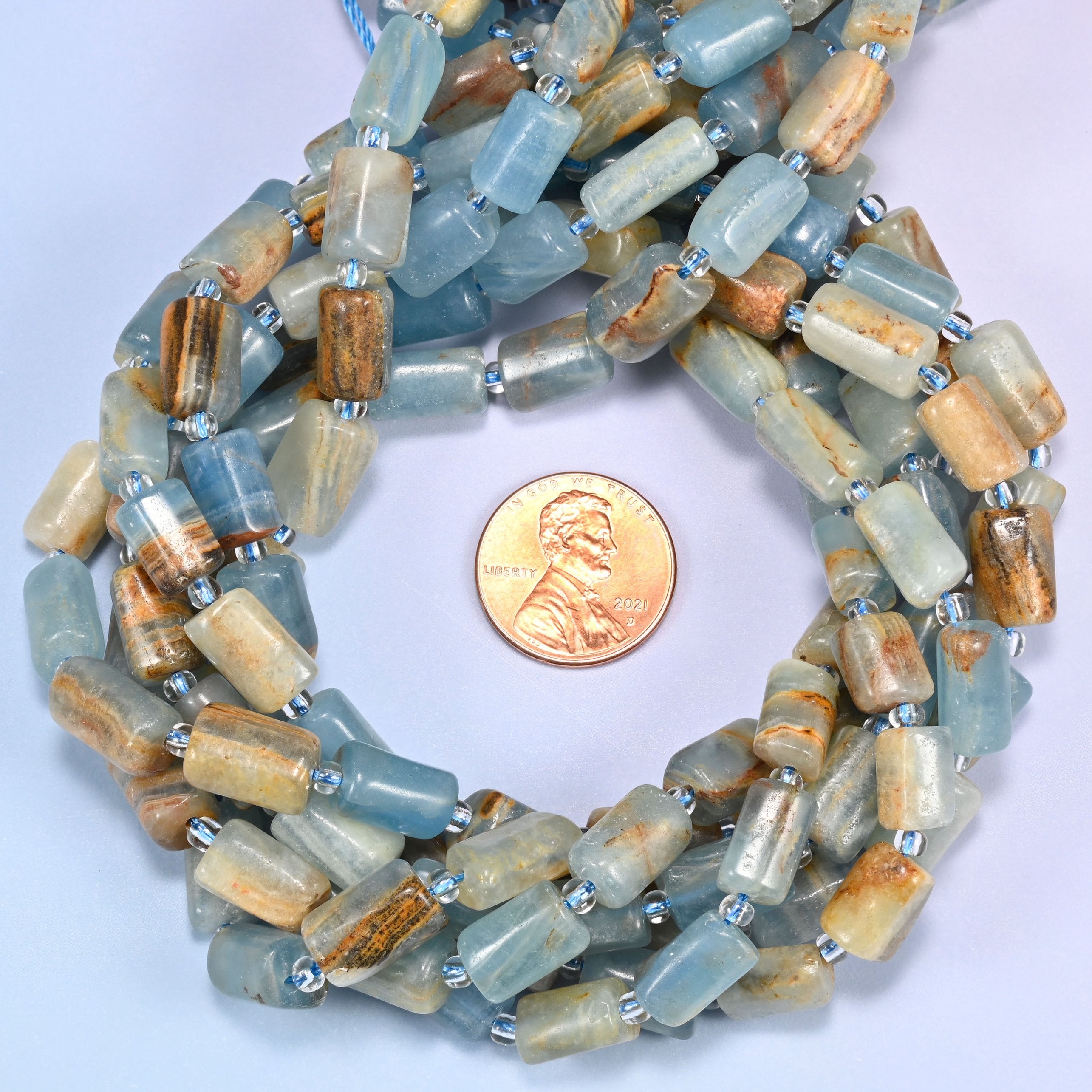 Multiple strands of faceted Blue Calcite tube beads gathered together on a lavender surface, loose ends visible with glass spacers.