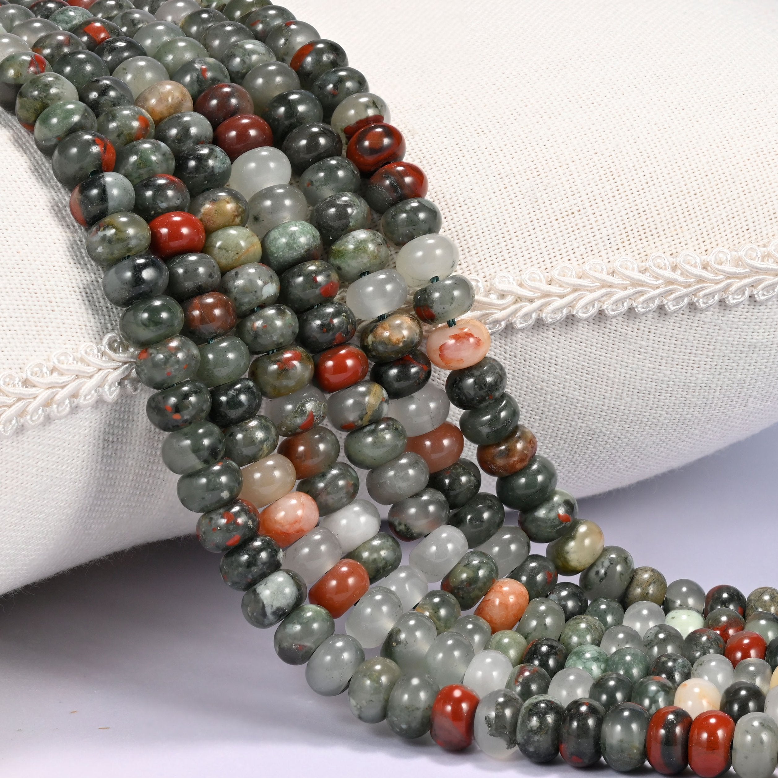 Close-up of multiple strands of polished African Bloodstone round beads in gray-green and red tones, draped over a cream fabric jewelry display bust.