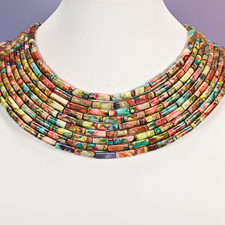 Multi-strand jasper tube bead necklace laid flat on a mannequin neck form against a lavender background, showing the full collar of layered colorful strands.