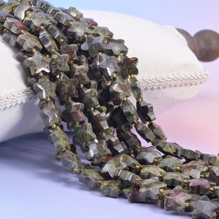 Dragon bloodstone faceted star beads over a white cushion, green gray stone with dark red patches.