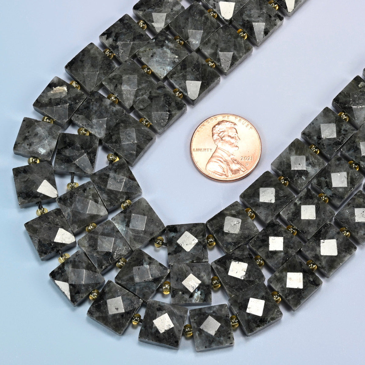 Close view of natural black labradorite faceted flat square strands curving beside a US penny for scale