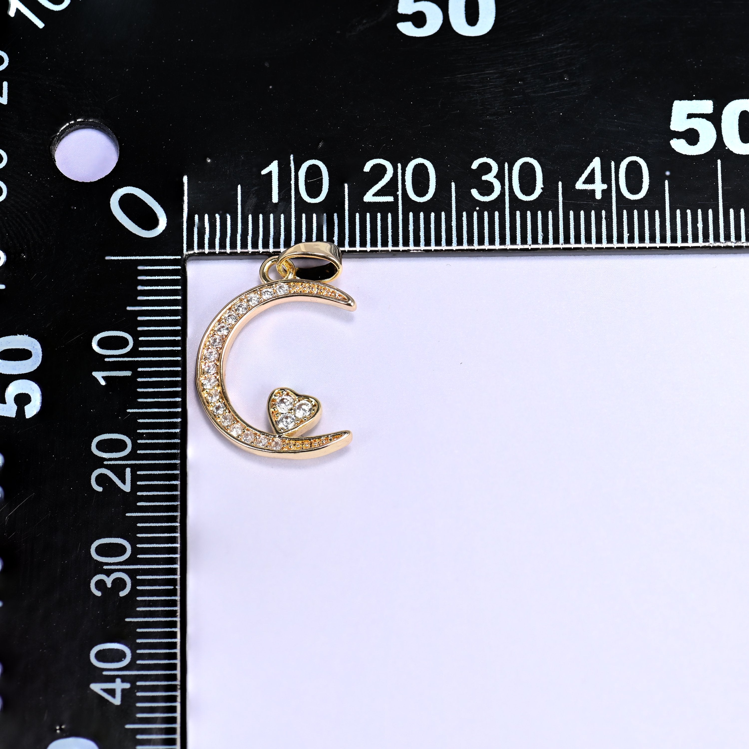 Gold moon heart zirconia charm on a black millimetre ruler, the crescent measuring roughly 20mm across.