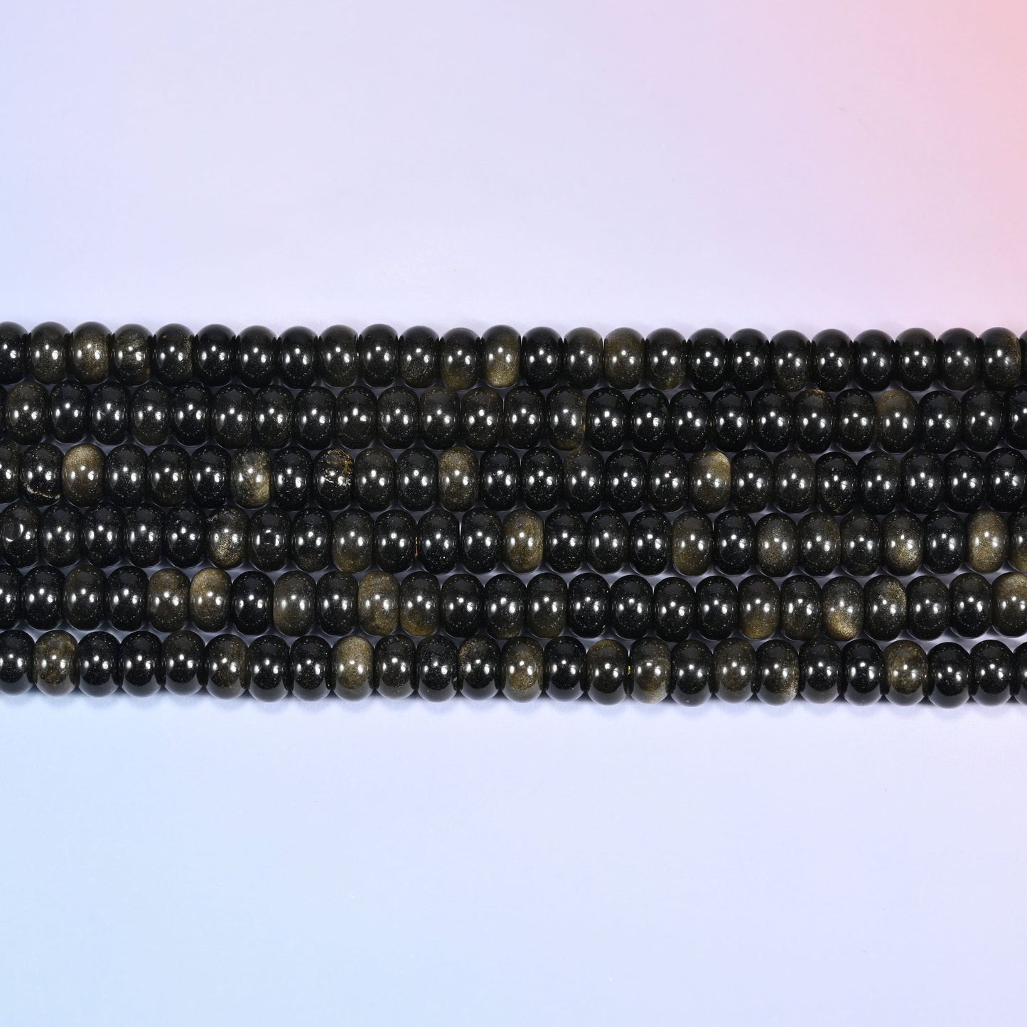 Overhead view of several rows of Gold Obsidian rondelle beads showing the black and golden shimmer on a light lavender surface.