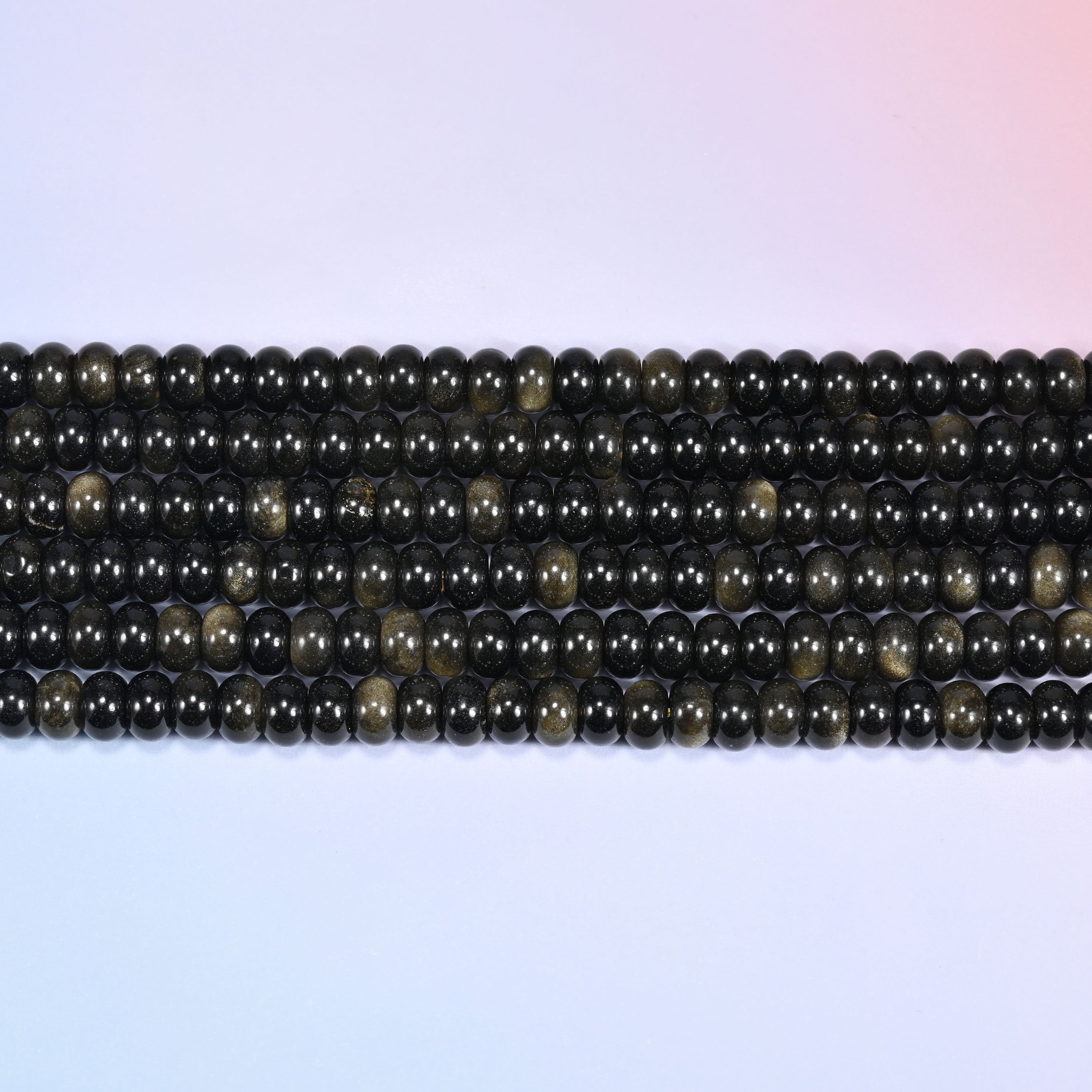 Overhead view of several rows of Gold Obsidian rondelle beads showing the black and golden shimmer on a light lavender surface.