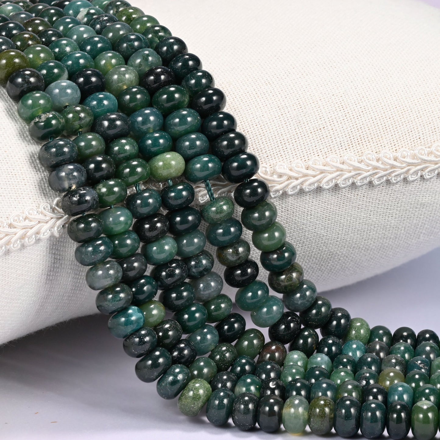 Close-up of multiple strands of polished Moss Agate rondelle beads in translucent green tones, draped over a cream fabric jewelry display bust.
