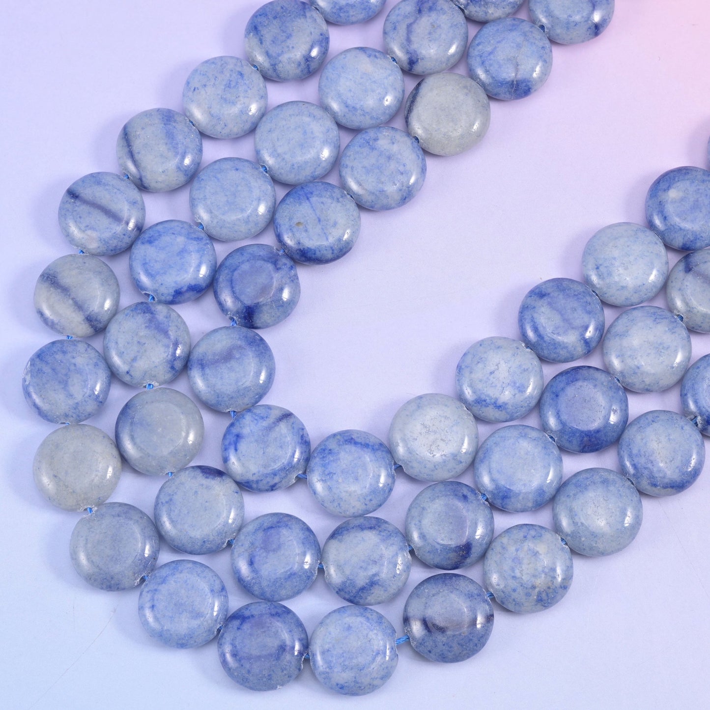 Two blue aventurine strands angling across lilac, showing the flat coin profile and softly rounded edges