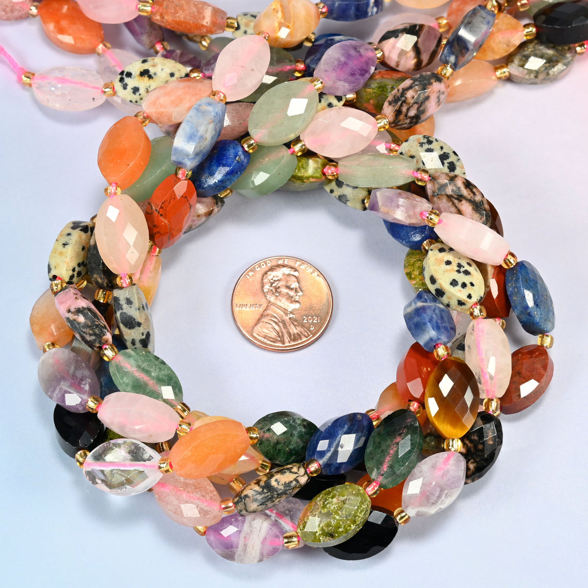Assorted gemstone faceted flat oval beads 9x14mm coiled in a loop around a US penny for scale, close-up of the faceted flat oval shape