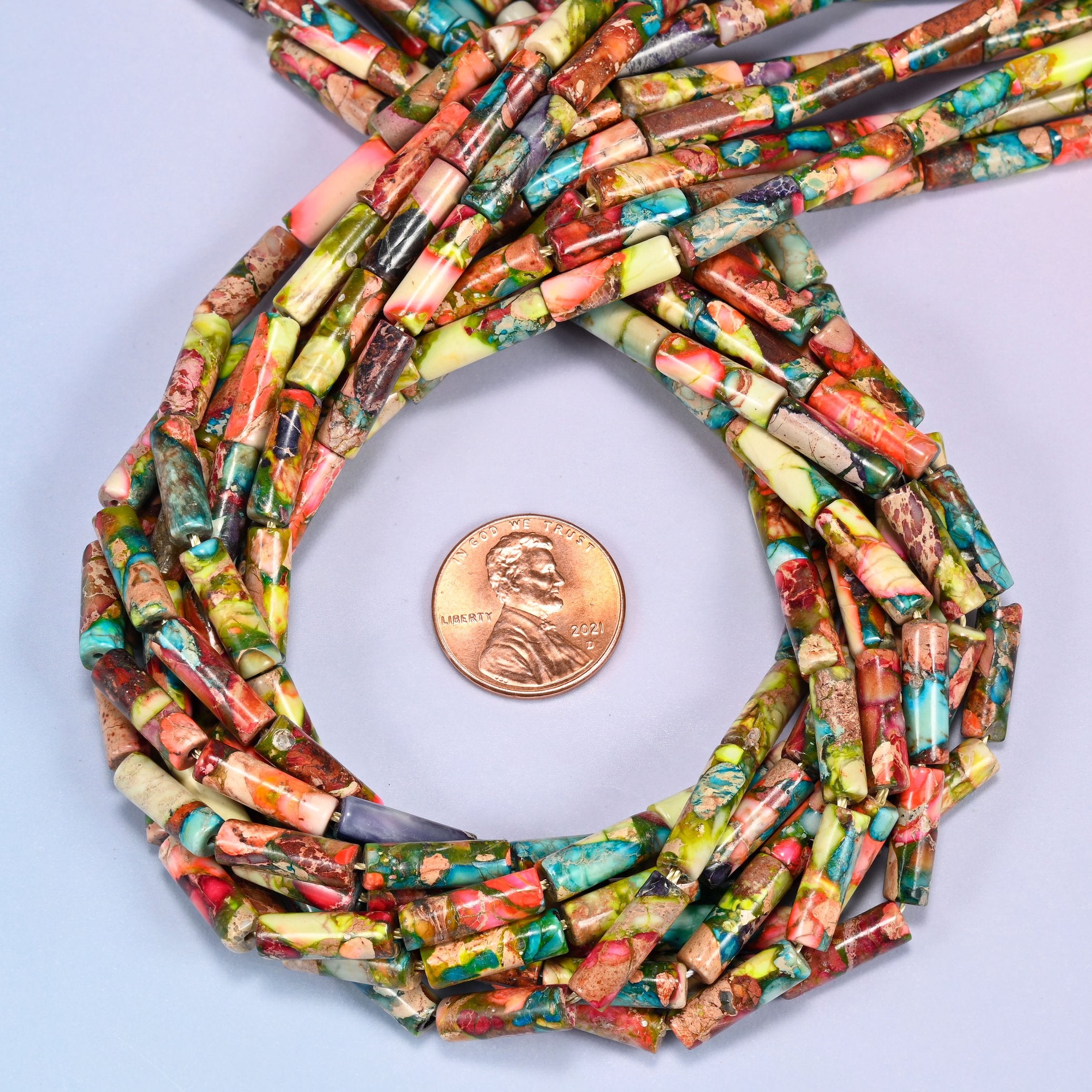Bundled coil of colorful jasper tube beads on a lavender background with a penny placed at the center for scale.