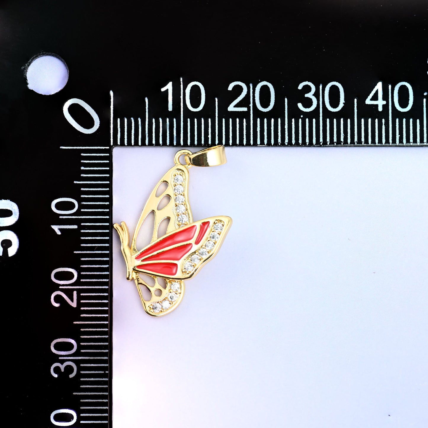 Flying butterfly charm pendant on a black millimetre ruler, the red enamel charm hanging upright from the zero mark for size reference.
