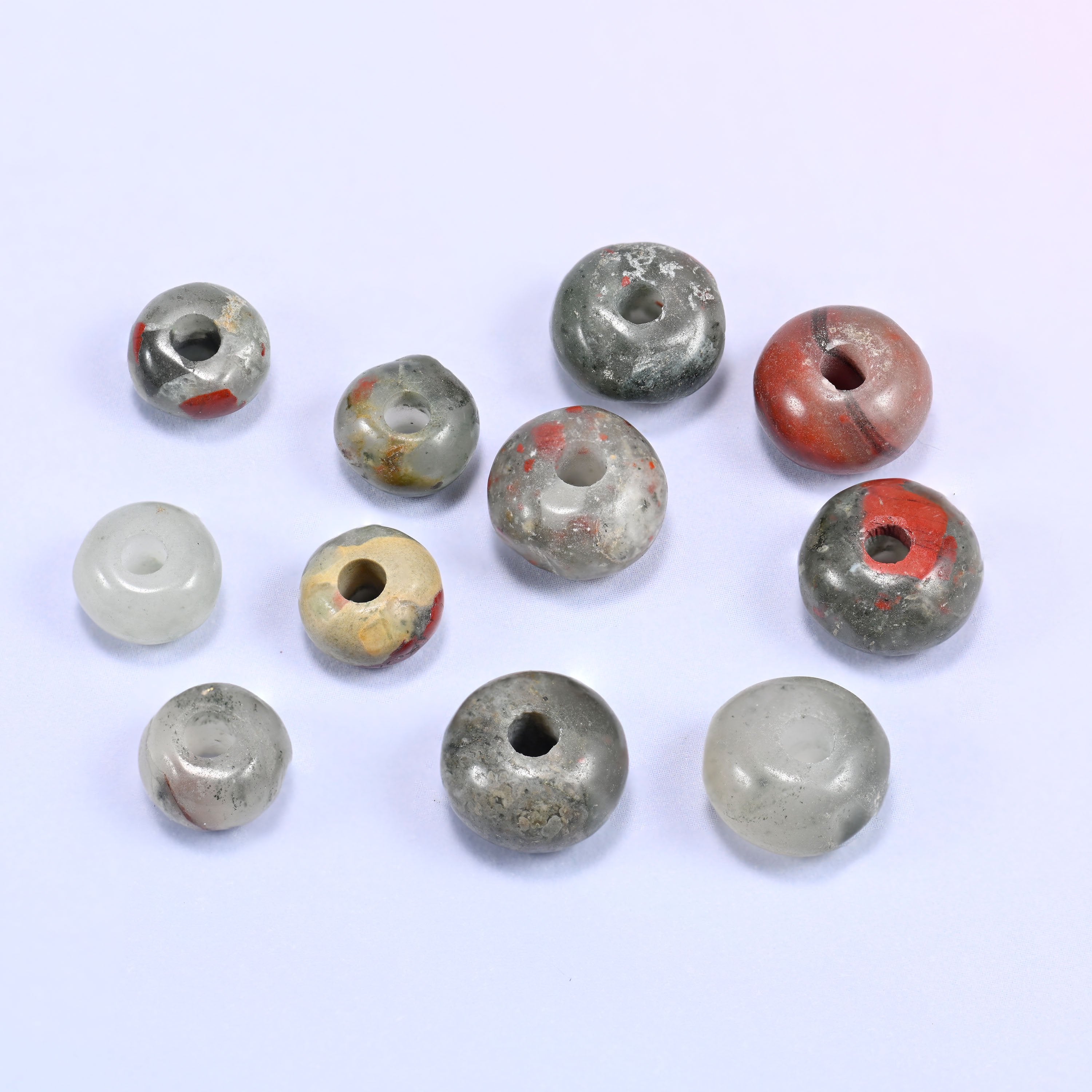 Close-up of individual loose African Bloodstone beads laid out on a white surface, showing the large center-drilled hole and varied coloring of each bead.