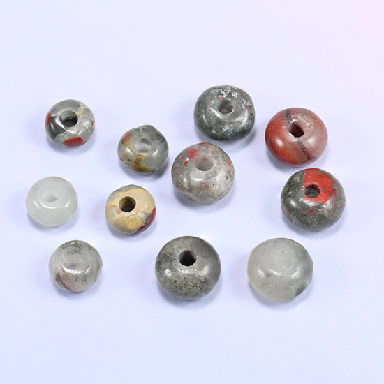 Close-up of individual loose African Bloodstone beads laid out on a white surface, showing the large center-drilled hole and varied coloring of each bead.