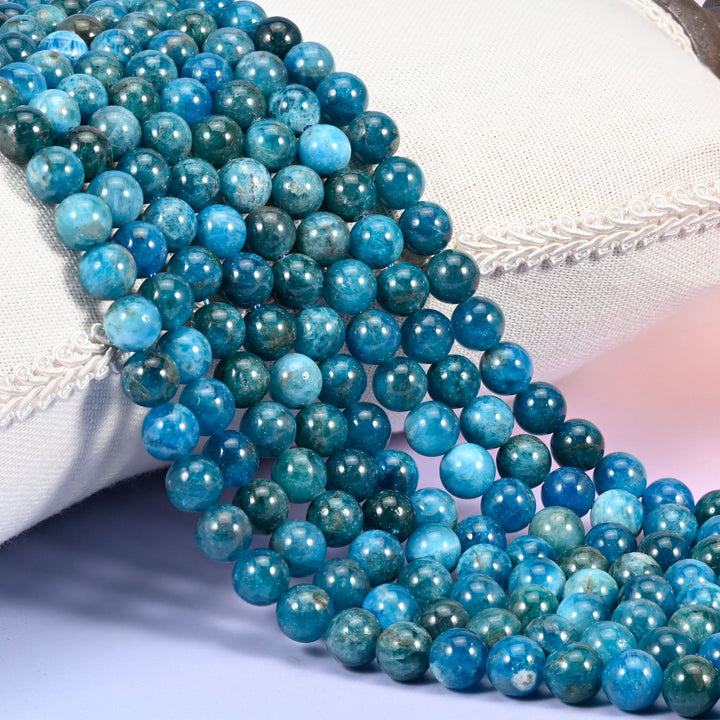 Blue Apatite smooth round beads, strands draped over a white cushion, deep teal rounds mottled with grey-green and lighter sky blue