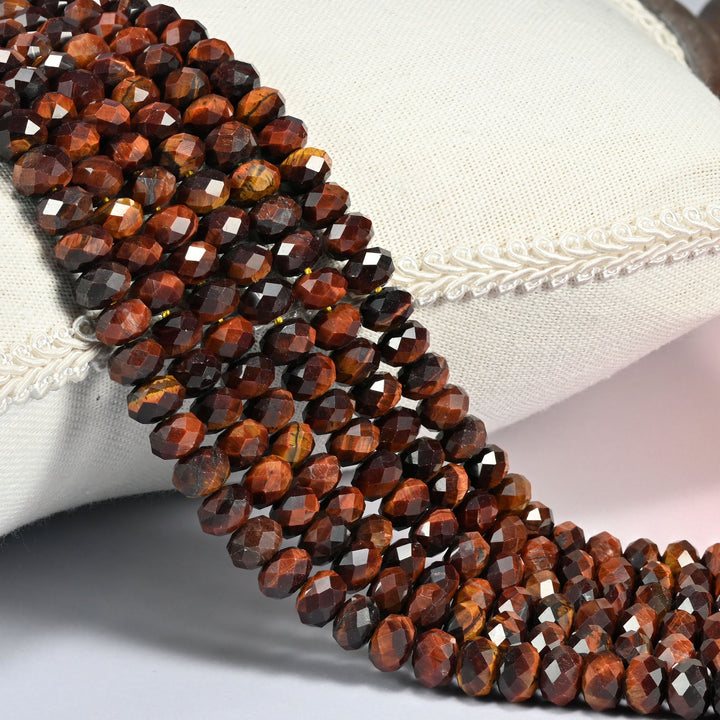 Red tiger eye faceted rondelle beads in deep red brown banded with near black, three strands draped over a cream linen cushion with braided trim