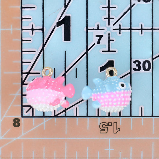 Enamel blowfish charms in pink and light blue placed side by side on a ruler for size comparison.