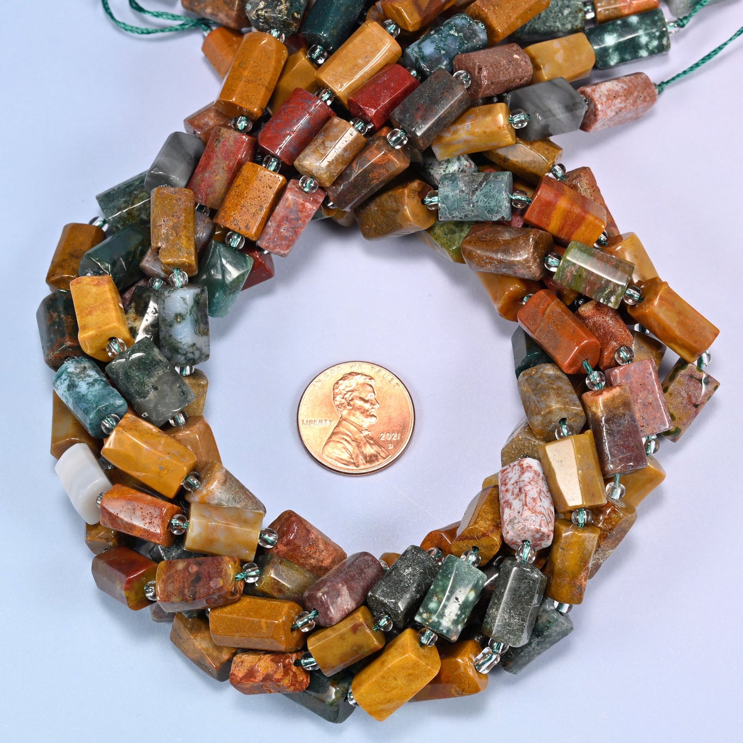 Multiple strands of faceted Ocean Jasper tube beads gathered together on a lavender surface, loose ends visible with glass spacers.