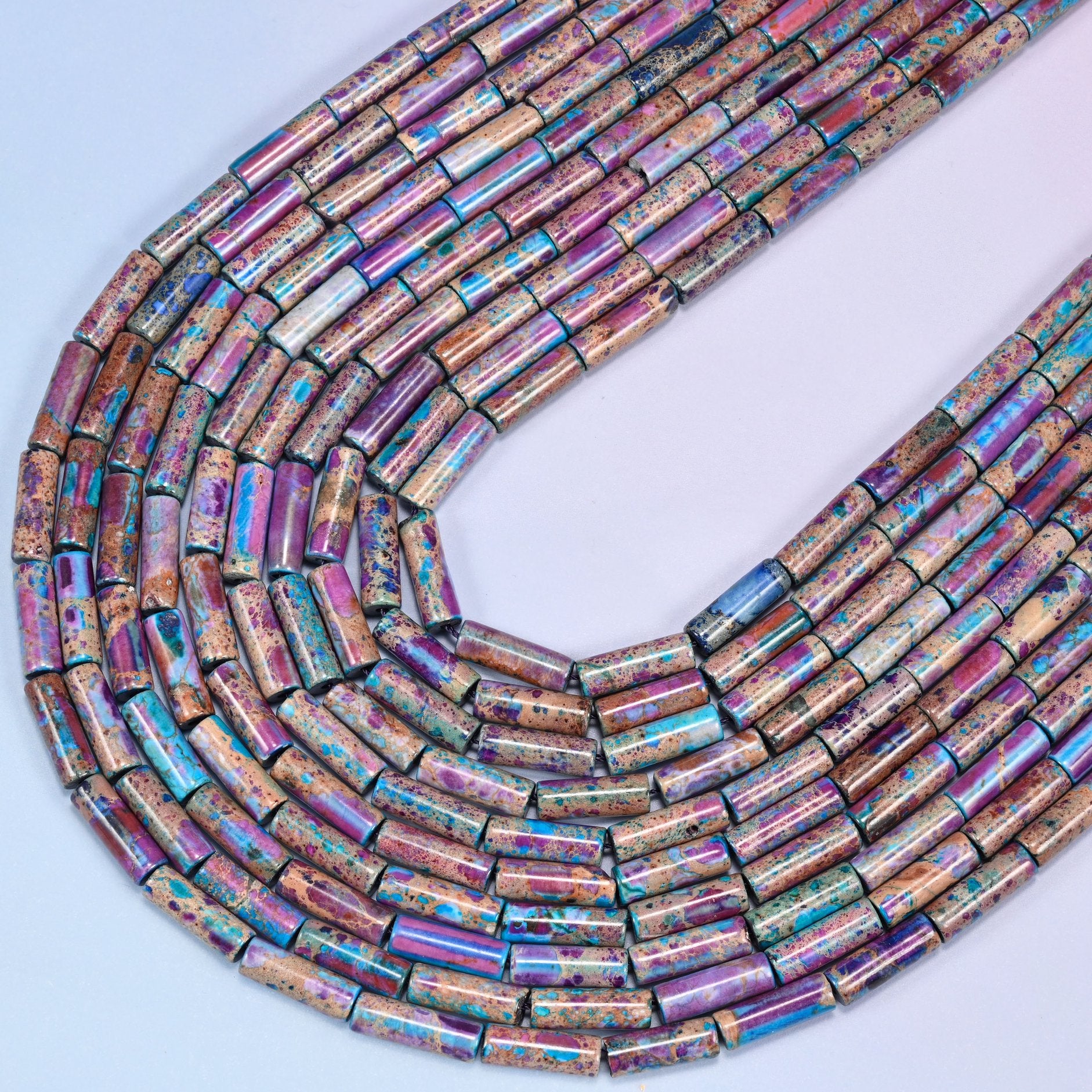 Close-up of purple and turquoise jasper tube beads coiled in a circular arrangement on a lavender background.