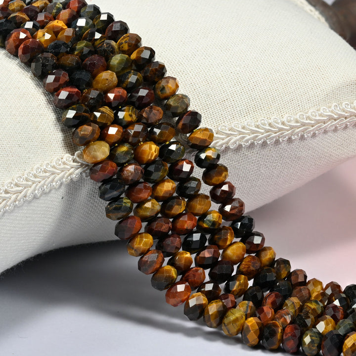 Tricolor tiger eye faceted rondelle beads 5x8mm in golden honey, brick red and blue-gray, several strands draped over a white cushion