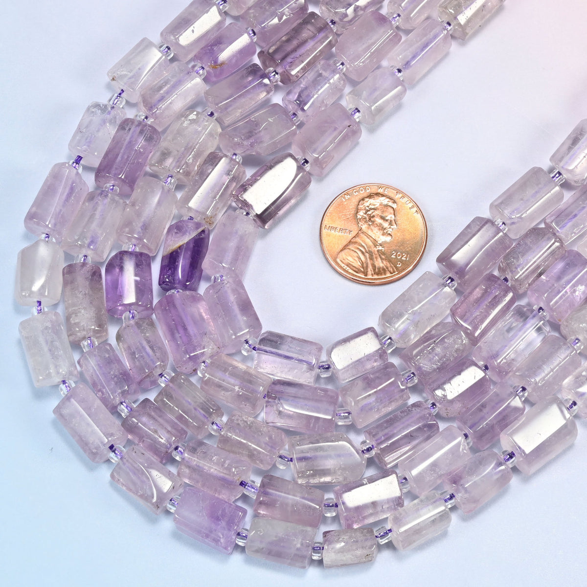 Lavender amethyst faceted tube beads coiled in a loop with a US penny resting at the right edge for scale.