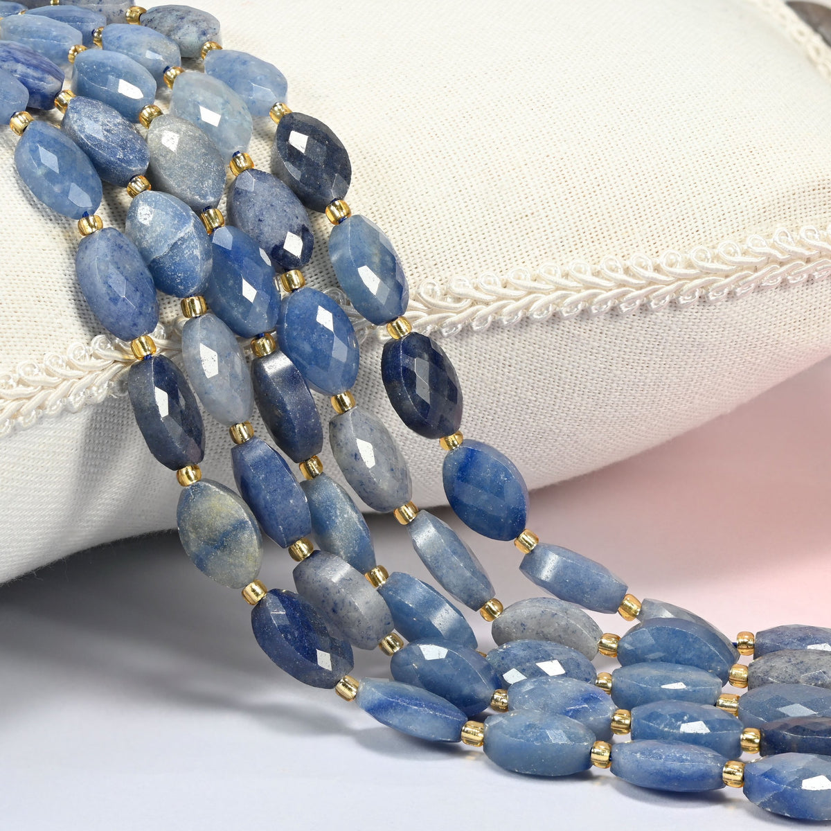 Blue aventurine faceted flat oval beads 9x14mm with small gold seed bead spacers, several strands draped over a white cushion