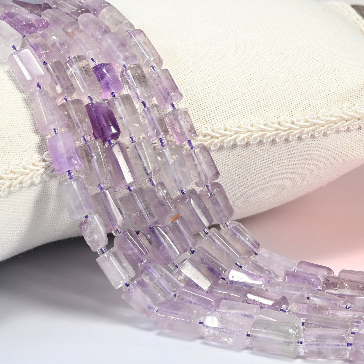 Lavender amethyst faceted tube beads draped in several strands over a white linen cushion with braided trim, soft lilac stone.