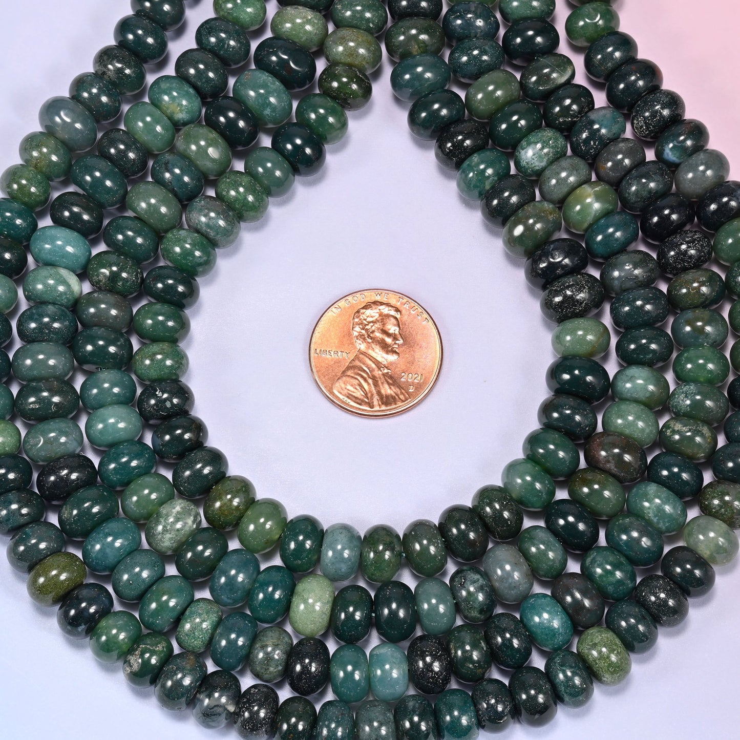 Bundled coil of Moss Agate rondelle beads on a lavender background with a penny placed at the center for scale.